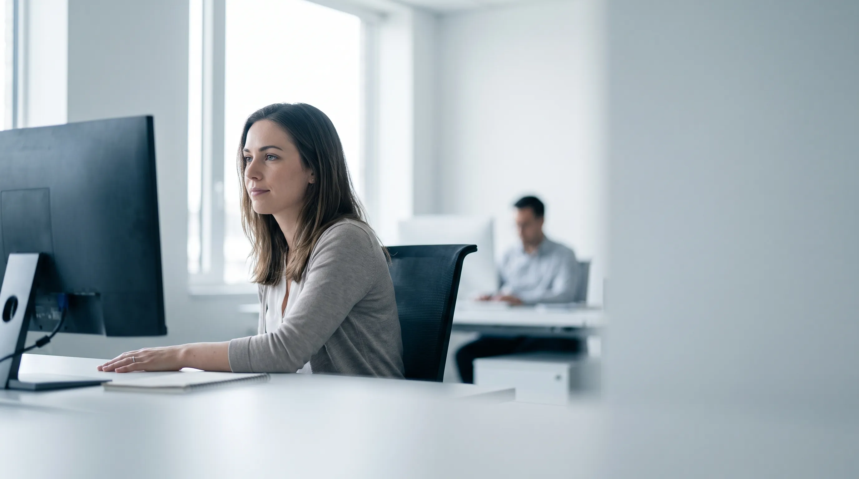Eine neue Mitarbeiterin arbeitet selbstständig an ihrem Schreibtisch in einem hellen Büro, im Hintergrund ein Kollege an einem eigenen Platz